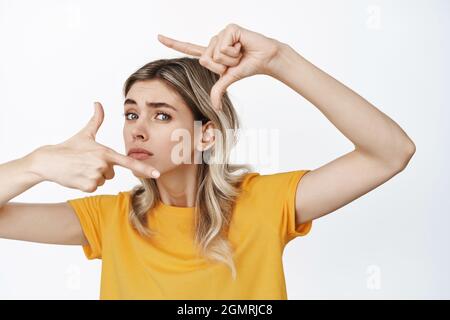 Lustige junge Frau, die durch Handrahmen mit unsicherem Gesichtsausdruck schaut, den perfekten Winkel für ein Foto sucht, etwas misst, über Weiß steht Stockfoto