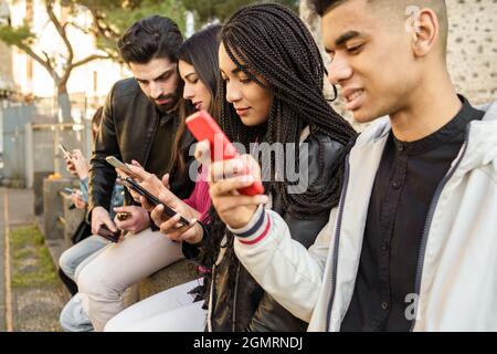 Generation z junge Menschen süchtig nach Smartphone und neuer Technologie. Gruppe von jungen Menschen mit gemischter Gruppe, die im Freien mit einem Mobiltelefon in den Händen sitzen. Te Stockfoto