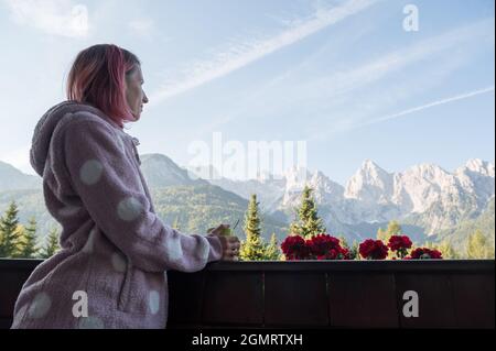 Junge Frau mit rosafarbenem Haar in kuscheligem, warmem Pullover, der sich an einem hölzernen Balkonzaun lehnt, eine Tasse Kaffee hält und einen wunderschönen Blick auf die Berge genießt. Stockfoto
