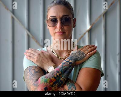 Schöne selbstbewusste gesunde junge kaukasische amerikanische Frau mit Sonnenbrille kreuzt ihre Arme und zeigt ihre feinen künstlerischen bunten Arm-Tattoos. Stockfoto