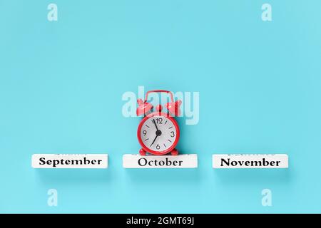 Holzkalender Herbstmonate und roter Wecker über Oktober auf blauem Hintergrund. Konzept Oktober Zeit Kreativ Draufsicht Flat Lay Minimal Style Copy Stockfoto