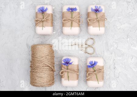 Verpackung von natürlicher handgemachter Seife, dekoriertes Kraftpapier, blaue Blume, Garnklee und Schere. Konzept der Bio-Kosmetik. Draufsicht Flat Lay Stockfoto