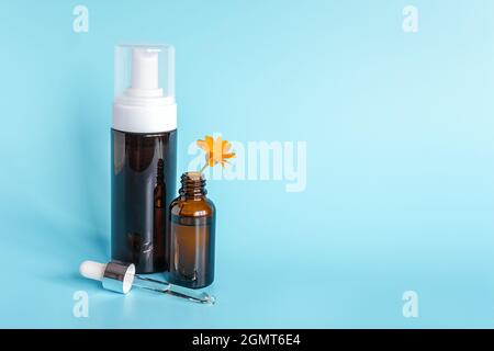Ätherisches Öl in offener brauner Tropfflasche mit liegender Glaspipette, große Flasche mit weißem Spender und orangefarbener Blütenkalendula auf blauem Hintergrund. C Stockfoto