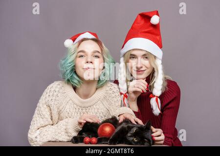 Junge süße Mädchen in weihnachtsmann Hut auf einem grauen Hintergrund, lustig lächelnd, halten Sie eine schwarze Katze. Mädchen mit grünen Haaren und blond. Zwei echte Freundinnen und ihr Lieblingstier Stockfoto