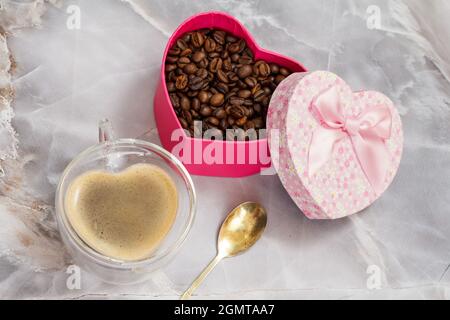 Eine Tasse Kaffee aus Glas und eine Geschenkbox mit gerösteten Kaffeebohnen auf dem Küchentisch. Die Tasse und die Box in Form eines Herzens. Draufsicht. Stockfoto