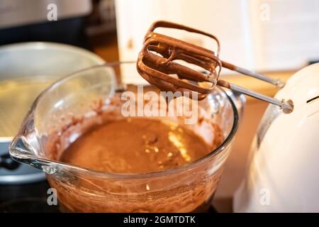 Schokoladenteig tropft vom Handmixer in eine Glasmischschüssel mit Backform und Kochbuch im Hintergrund. Stockfoto