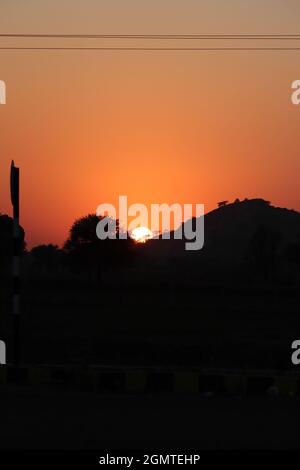 Blick auf volle runde gelbe helle Sonne und gedämpftes Sonnenlicht, Sonnenuntergang hinter Silhouette wilden Bäumen und kleinen Hügel, die volle Helligkeit und scharf der Sonne in Stockfoto