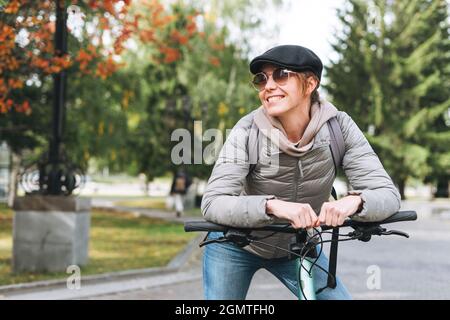 Porträt einer modischen jungen hübschen Frau mit Mütze und Sonnenbrille auf dem Fahrrad am sonnigen Herbsttag im Stadtpark Stockfoto