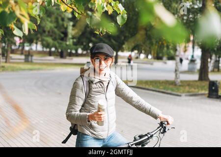 Porträt einer modischen jungen hübschen Frau in Mütze und Sonnenbrille auf dem Fahrrad mit Thermobecher am sonnigen Herbsttag in der Stadt Stockfoto