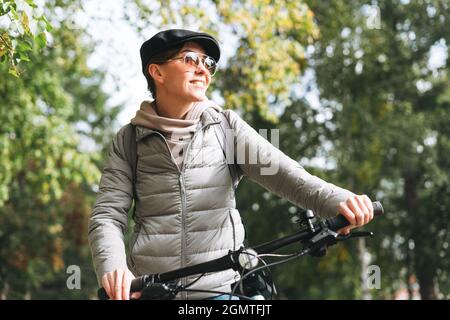 Porträt einer modischen jungen hübschen Frau mit Mütze und Sonnenbrille auf dem Fahrrad am sonnigen Herbsttag in der Stadt Stockfoto