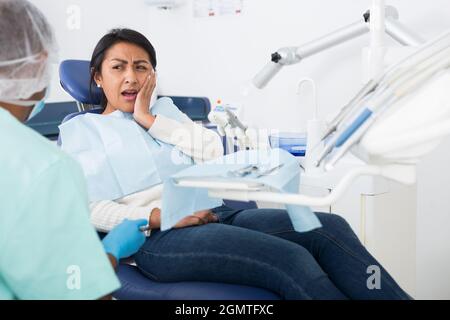 Latina, die an Zahnschmerzen leidet, spricht mit einem Stomatologen, während sie in der Klinik auf einem Zahnstuhl sitzt Stockfoto