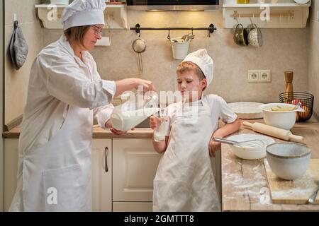 Ein Junge in der Kleidung des Küchenchefs trinkt Milch aus einem Glas, während er in der Küche einen Apfelkuchen kocht Stockfoto