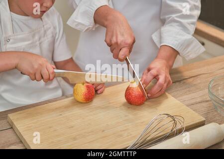 Mutter und Sohn in weißen Chefkleidern schnitten in ihrer eigenen Küche mit einem Messer Birnen. Eine Erwachsene Frau mit einem kleinen Jungen wird einen Kuchen kochen Stockfoto