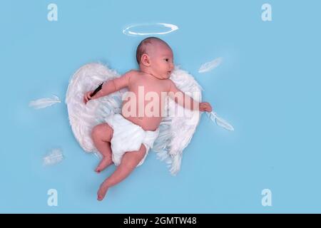 Ein neugeborener Junge mit weißen Engelsflügeln auf blauem Studiohintergrund, Kopierraum. Ein kaukasisches Kleinkind in einer Windel im Alter von einem Monat Stockfoto