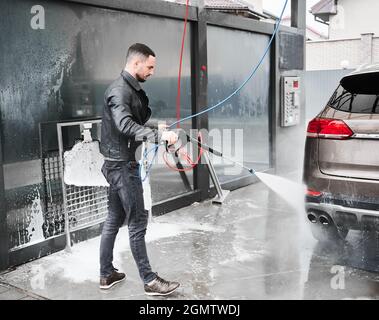 Junger Mann, der im Freien an der Autowaschanlage Auto waschen kann. Hübscher Arbeiter, der Auto putzt, mit Hochdruckwasser. Stockfoto