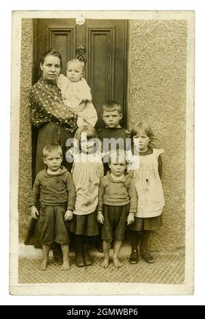 Ursprüngliche, sehr klare, edwardianische Porträtpostkarte mit einer typischen armen Arbeitermutter mit einer großen Familie von sechs ernst aussehenden kleinen Kindern und einem Baby in ihren Armen, einige Kinder sind barfuß, tragen schmuddelige, zerrissene Kleidung. Die Mädchen sind in Schürzkügelchen gekleidet, Jungen tragen knabberige Pullover, vor der Haustür, aus dem Studio von J.H. Ormerod, Seacombe, Wallasey, The Wirral, England, Großbritannien um 1907 Stockfoto
