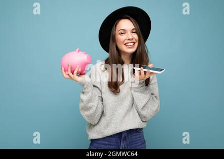 Portrait von glücklich positiv lächelnd lachend junge schöne Brünette Frau trägt stilvolle Pullover und schwarzen Hut isoliert auf blauem Hintergrund mit Stockfoto