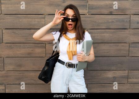 Faszinierende schöne lächelnde charmante junge Brunet Mädchen Blick auf Kamera hält Computer Laptop und schwarze Sonnenbrille in weißem T-Shirt gelb Stockfoto