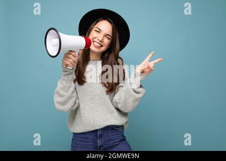 Porträt von jungen schönen positiv lächelnden glücklich Brünette Frau mit aufrichtigen Emotionen tragen trendigen schwarzen Hut und grauen Pullover isoliert über blau Stockfoto