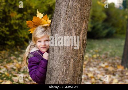 Schönes Mädchen mit orangefarbenen Blättern im Haar versteckt hinter Baumstamm Stockfoto