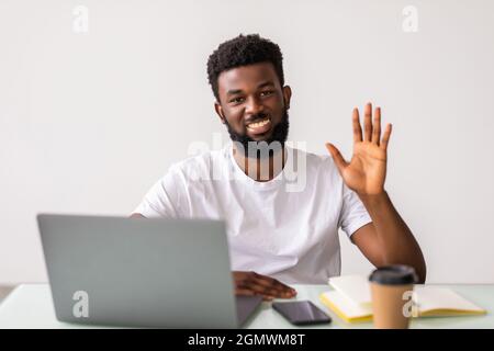 Glücklicher schwarzer Mann an seinem Schreibtisch zu Hause, der Hallo sagte und die Hand an die Kamera schwenkte. Web Cam Laptop Computer Bildschirm Ansicht von Studenten für Bildungs-Web Stockfoto