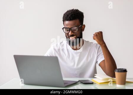 Glücklicher Geschäftsmann feiert, gewinnt. Gewinner, schwarzer Mann in der Freizeit im Büro mit Laptop Stockfoto