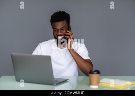Fröhlicher afro-amerikanischer Kerl macht Multi-Aufgaben Arbeit, ein gemischter Mann tragen smart Casual tragen tragen, sprechen am Telefon, Notizen und mit einem Schoß Stockfoto
