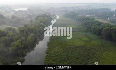 Nebliger Morgen über einer Ebene und Flussauen Wiese in der Nähe eines ländlichen Dorfes mit einem Haus. Ländliche Landschaft im Tal aus der Höhe des Fluges Stockfoto