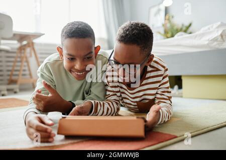 Porträt von zwei afroamerikanischen Jungen im Teenageralter, die zusammen ein digitales Tablet benutzen, während sie auf dem Boden in einem gemütlichen Raum liegen und Platz kopieren Stockfoto