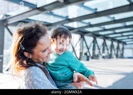 Ein Jahr altes Baby mit schelmischen Blick lächelnd von seiner tausendjährigen Mutter auf der Straße gefangen Stockfoto