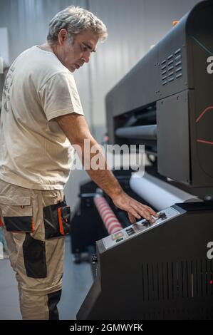 Der Bediener des Technikers wechselt die Papierrolle auf einem großen hochwertigen Industriedrucker und Plottergerät im digitalen Druckereibüro Stockfoto