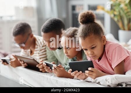 Vier afroamerikanische Kinder, die Gadgets in Reihe benutzen, während sie zusammen auf dem Bett liegen, Konzept mehrerer Geschwister Stockfoto