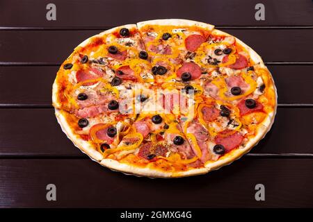 Auf einem Pappteller auf einem Holztisch wird eine ganze rustikale Pizza capriciosa mit Würstchen, Speck, Pfeffer und Pilzen serviert Stockfoto