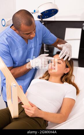 Junge hispanische Frau, die in den Spiegel schaut, während professionelle männliche Kosmetikerin sie vor der Gesichtsbehandlung im kosmetologischen Büro konsultiert. Stockfoto