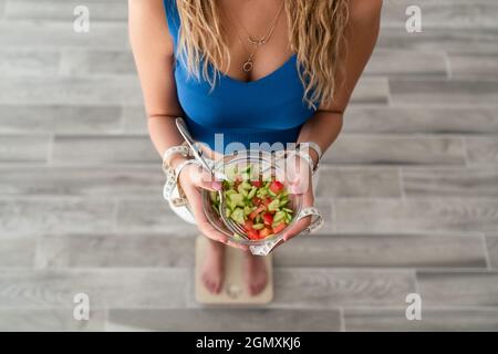 Junge Frau isst Salat für ein gesundes Leben und einen gesunden Körper. Hält ein Maßband, um ihre Taille zu messen und steht auf einer Waage. Stockfoto