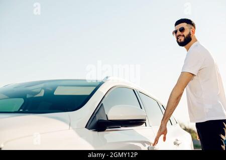 Selbstbewusster junger Geschäftsmann mit Sonnenbrille, Mann öffnet die Tür seines Autos, Mann sitzt in seinem Auto im Freien in der Stadt Stockfoto