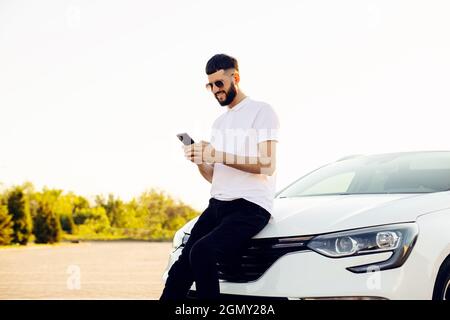 Hübscher junger Mann mit Smartphone in der Nähe eines modernen Autos im Freien, hübscher Mann auf dem Hintergrund eines Autos Stockfoto