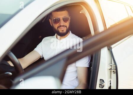 Der glückliche Besitzer, ein hübscher bärtiger junger Mann, in weißem T-Shirt, sitzt entspannt in seinem neuen Auto und blickt freudig aus dem Fenster Stockfoto