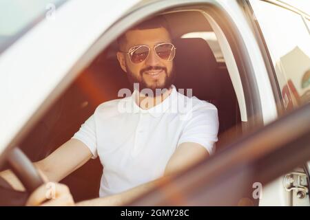 Der glückliche Besitzer, ein hübscher bärtiger junger Mann, in weißem T-Shirt, sitzt entspannt in seinem neuen Auto und blickt freudig aus dem Fenster Stockfoto