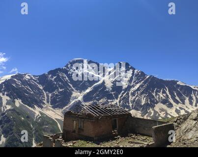 Altes verlassenes Haus in den Bergen. Ruinen einer Hütte ohne Dach auf dem Hintergrund der schneebedeckten Gipfel der Berge. Stockfoto