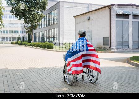 Rückansicht eines jungen Veteranen, bedeckt mit der usa-Flagge, der im Rollstuhl in der Stadt sitzt Stockfoto