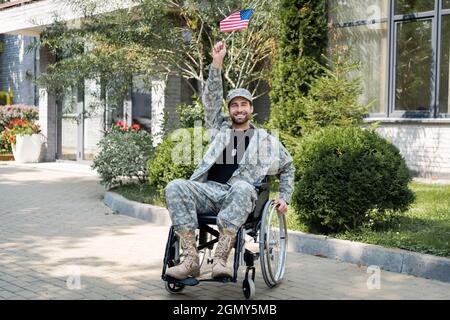 Junger und glücklicher behinderter Veteran, der im Rollstuhl sitzt, mit einer kleinen us-Flagge in erhobener Hand Stockfoto