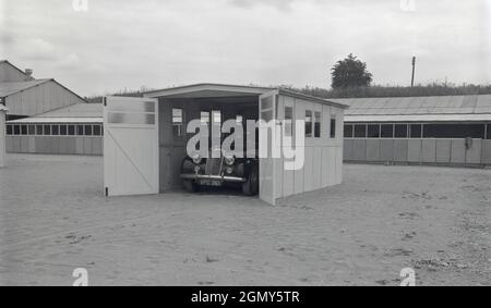 1950er Jahre, historisch, draußen auf einem breiten, Ebenen Gebiet in einem leichten Industriegebiet, ein Auto der damaligen Zeit, geparkt in einer Beton-, Fertiggarage, vielleicht dort zu Demonstrationszwecken gebaut, Witney, Oxford, England, Großbritannien. Diese Garage oder der Motorschutz wurde aus Betonfertigteilen mit Dach und Holztüren gebaut, die in Abschnitte geschraubt wurden. Mit der Zunahme des Fahrzeugbesitzes in den 1950er und 1960er Jahren wurden diese Selbstbau- oder Betonwerkzeugaragen in Großbritannien populär, oft in kleinen Anzeigen in Zeitungen und Zeitschriften gesehen, die als DIY-Projekt für den „praktischen Haushälter“ beworben wurden. Stockfoto