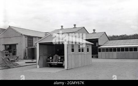 1950er Jahre, historisch, draußen auf einem breiten, Ebenen Gebiet in einem leichten Industriegebiet, ein Auto der damaligen Zeit, geparkt in einer Beton-, Fertiggarage, vielleicht dort zu Demonstrationszwecken gebaut, Witney, Oxford, England, Großbritannien. Diese Garagen oder Motorschutzräume wurden aus Betonfertigteilen hergestellt, die in Abschnitte miteinander verschraubt wurden, mit einem Welldach und einer Metallaufrolltür. Mit der Zunahme des Fahrzeugbesitzes in den 1950er und 1960er Jahren wurden diese Selbstbau- oder Betonfertigteilwerkhäuser in Großbritannien populär, was oft in kleinen Anzeigen in Zeitungen und Zeitschriften für den „praktischen Hausbesitzer“ zu sehen ist. Stockfoto