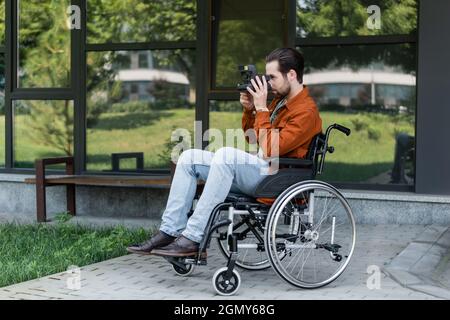 Junger behinderter Mann im Rollstuhl, der in der Nähe eines Gebäudes mit Glasfassade Fotos mit einer Vintage-Kamera gemacht hat Stockfoto