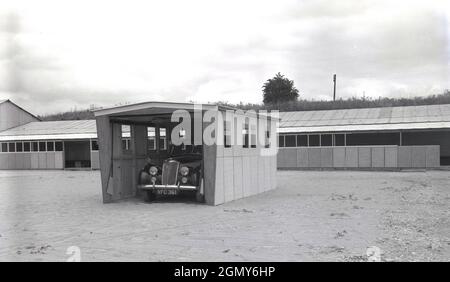 1950er Jahre, historisch, draußen auf einem breiten, Ebenen Gebiet in einem leichten Industriegebiet, ein Auto der damaligen Zeit, geparkt in einer Beton-, Fertiggarage, vielleicht dort zu Demonstrationszwecken gebaut, Witney, Oxford, England, Großbritannien. Diese Garagen oder Motorschutzräume wurden aus Betonfertigteilen hergestellt, die in Abschnitte miteinander verschraubt wurden, mit einem Dach und einem Metallaufrolltor oder über der Tür. Mit der Zunahme des Fahrzeugbesitzes in den 1950er und 1960er Jahren wurden diese Selbstbau- oder Betonbausätze in Großbritannien populär, was oft in kleinen Anzeigen in Zeitungen und Zeitschriften zu sehen ist, die dem „praktischen Haushälter“ beworben wurden. Stockfoto