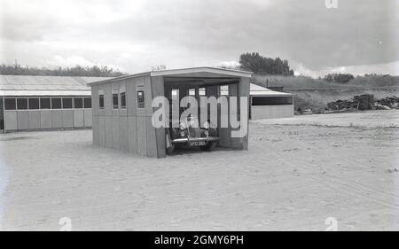 1950er Jahre, historisch, draußen auf einem breiten, Ebenen Gebiet in einem leichten Industriegebiet, ein Auto der damaligen Zeit, geparkt in einer Beton-, Fertiggarage, vielleicht dort zu Demonstrationszwecken gebaut, Witney, Oxford, England, Großbritannien. Diese Garagen oder Motorschutzräume wurden aus Betonfertigteilen hergestellt, die in Abschnitte miteinander verschraubt wurden, mit einem Dach und einem Metallaufrolltor oder über der Tür. Mit der Zunahme des Fahrzeugbesitzes in den 1950er und 1960er Jahren wurden diese Selbstbau- oder Betonfertigteilwerkhäuser in Großbritannien populär, was oft in kleinen Anzeigen in Zeitungen und Zeitschriften für den „praktischen Hausbesitzer“ zu sehen ist. Stockfoto