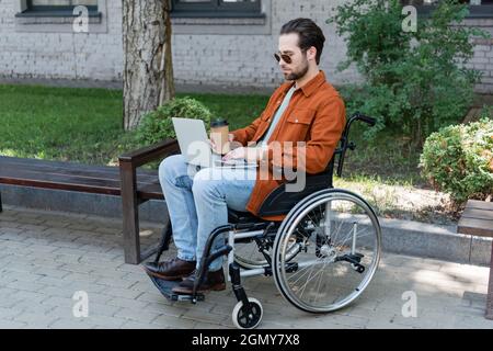 Behinderter Mann mit einem Getränk zum Mitnehmen, der im Rollstuhl sitzt und an einem Laptop arbeitet Stockfoto