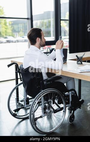 Ein behinderter Geschäftsmann im Rollstuhl, der im Büro in der Nähe eines Laptops auf dem Smartphone nachmittlungsbereit ist Stockfoto