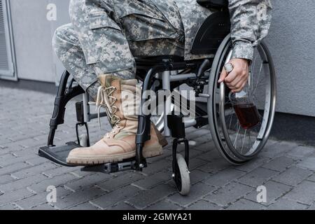 Beschnittene Ansicht eines behinderten Soldaten im Rollstuhl, der eine Flasche Whiskey im Freien hält Stockfoto